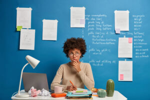 Thoughtful attractive woman in spectacles writes in diary to do list of goals, makes notes in personal organizer, poses at cozy coworking space, creats article on scientific topic, thinks at plans